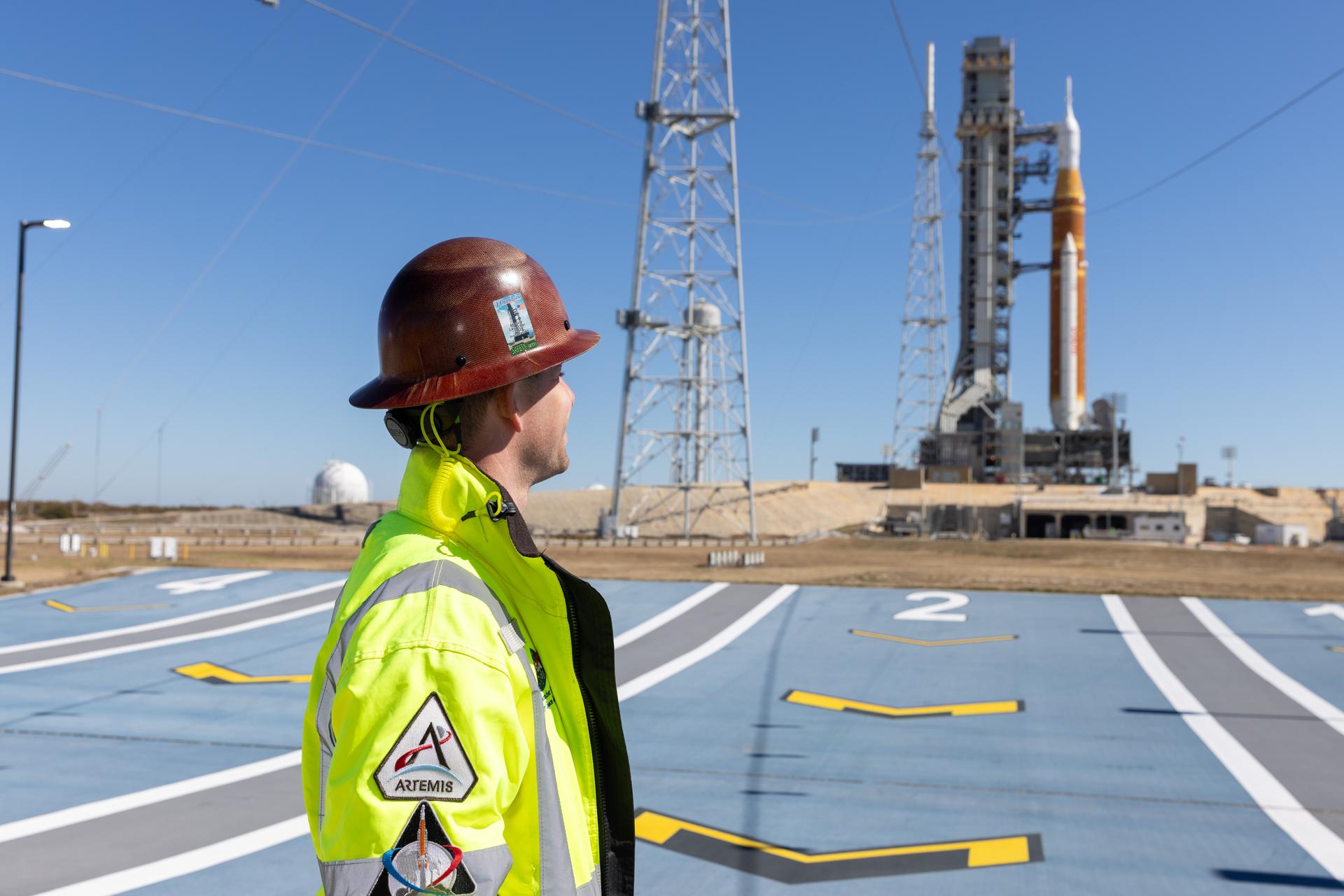 Image shows Jesse Berdis standing standing at the pad of Launch Complex 39B at NASA's Kennedy Space Center in Florida. Behind him are clear blue skies and NASA's SLS (Space Launch System) rocket and Orion spacecraft for the Artemis II mission. Photo credit: Kim Shiflett